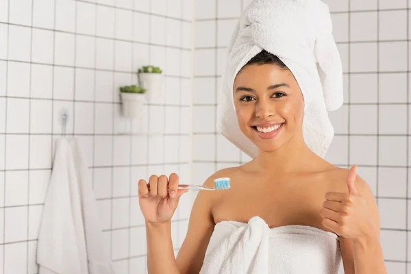 Giovane donna afroamericana avvolta in asciugamani in possesso di spazzolino da denti e mostrando gesto ok in bagno — Foto stock