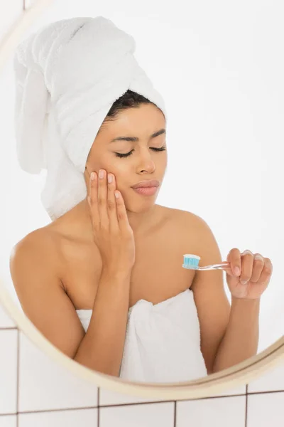 Jeune femme afro-américaine tenant une brosse à dents et souffrant de maux de dents avec les yeux fermés près du miroir dans la salle de bain — Photo de stock