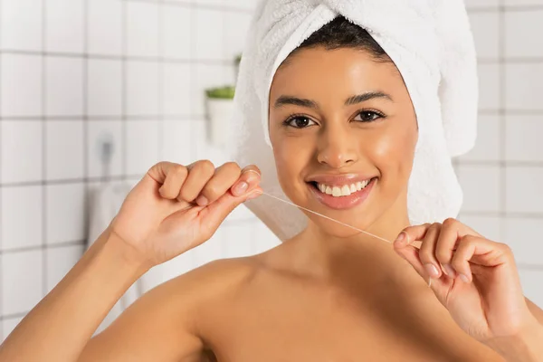 Sorridente jovem afro-americano mulher envolto em toalha segurando fio dental em mãos perto do rosto no banheiro — Fotografia de Stock