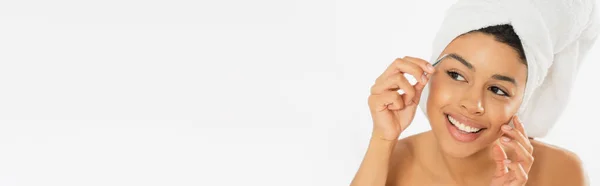 Mujer afroamericana joven y positiva arrancando cejas con pinzas sobre fondo blanco, pancarta - foto de stock