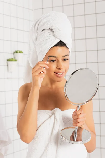 Positivo giovane donna afro-americana avvolto in asciugamani pizzicare le sopracciglia con pinzette in bagno — Foto stock