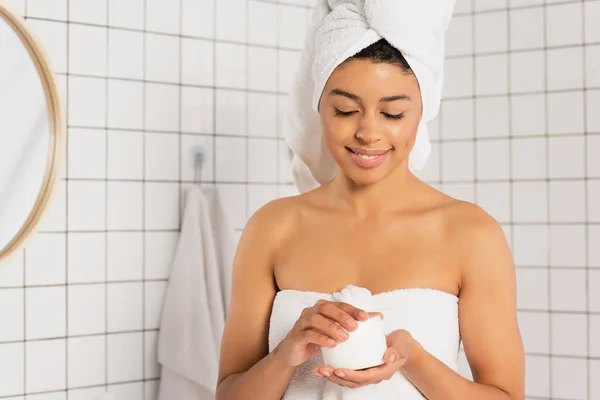 Sonriente joven afroamericana mujer sosteniendo tarro con crema en el baño - foto de stock