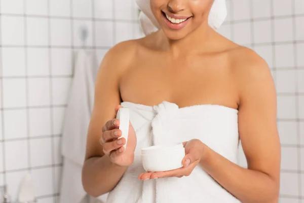 Vista parziale di giovane donna afroamericana avvolta in asciugamani apertura vaso crema in bagno — Foto stock