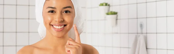 Mujer afroamericana joven aplicando crema en la cara con el dedo en el baño, pancarta - foto de stock