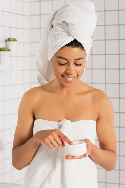 Jeune femme afro-américaine enveloppée dans des serviettes toucher la crème dans un bocal avec doigt dans la salle de bain — Photo de stock