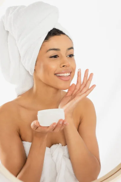Молодая африканская американка, завернутая в полотенца, нанося крем из банки на лицо на белом фоне — стоковое фото