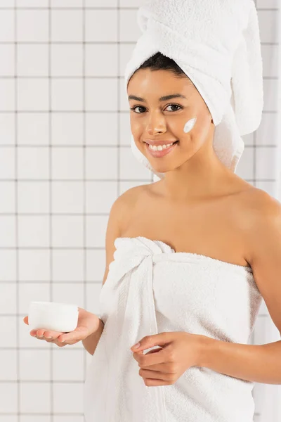 Sonriente joven afroamericana mujer sosteniendo tarro con crema sobre fondo blanco texturizado - foto de stock
