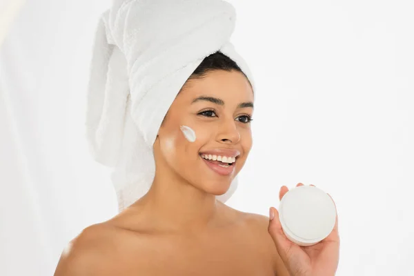 Souriant jeune femme afro-américaine présentant pot avec de la crème et regardant loin sur fond blanc — Photo de stock