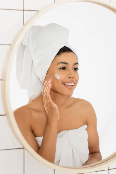 Jeune femme afro-américaine enveloppée dans des serviettes appliquer la crème sur le visage et regarder dans le miroir dans la salle de bain — Photo de stock