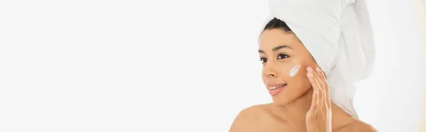 Улыбающаяся молодая африканская американка наносит крем на лицо на белом фоне, баннер — стоковое фото