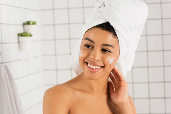 Jeune femme afro-américaine avec crème sur les joues touchant le cou dans la salle de bain — Photo de stock