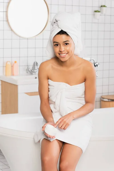 Sonriente joven afroamericana mujer sentada en la bañera y sosteniendo frasco con crema en el baño - foto de stock