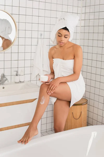 Jeune femme afro-américaine enveloppée dans des serviettes appliquer la crème sur la jambe dans la salle de bain — Photo de stock