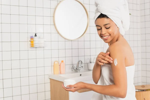 Sonriente joven afroamericana mujer aplicando crema en el brazo en el baño - foto de stock