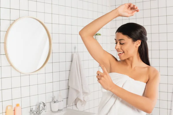 Jeune femme afro-américaine enveloppée dans une serviette de pulvérisation avec déodorant dans la salle de bain — Photo de stock