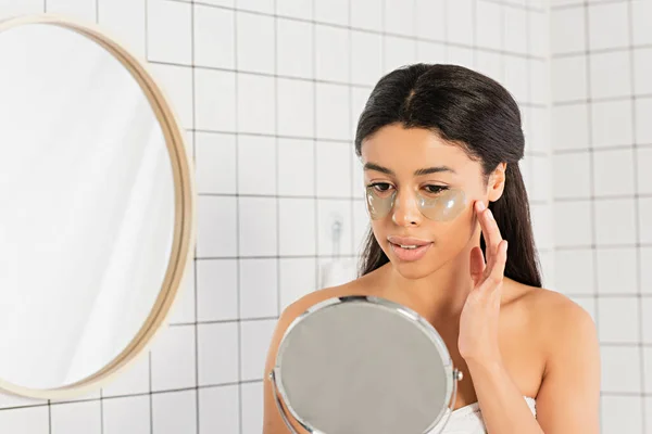 Enfocada joven afroamericana mujer agarrada de la mano cerca de la cara con parches en los ojos y mirando el espejo en el baño - foto de stock