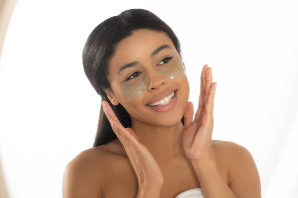 Happy young african american woman holding hands near face with eye patches in bathroom — Stock Photo