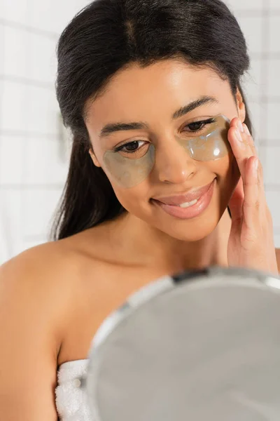 Jovem afro-americana segurando mão perto do rosto com manchas nos olhos e olhando para o espelho no banheiro — Fotografia de Stock