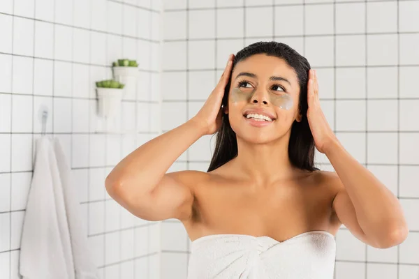 Joven afroamericana mujer envuelta en toalla con parches en los ojos tomados de la mano en la cabeza en el baño - foto de stock