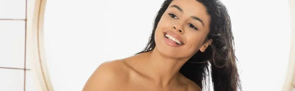 Sonriente joven afroamericana mujer con el pelo mojado mirando en el espejo en el baño, pancarta - foto de stock