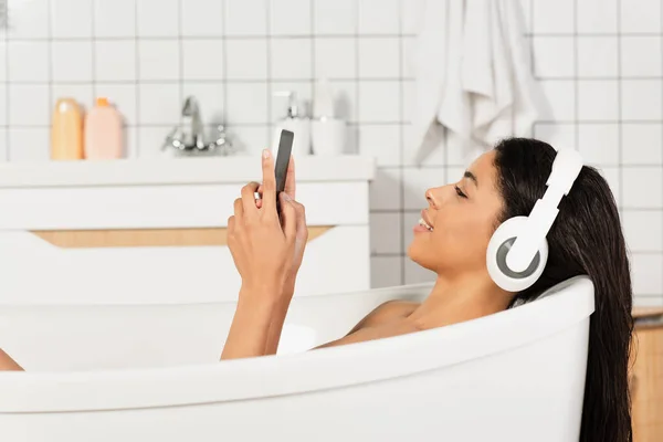 Vista lateral de la joven sonriente escuchando música en auriculares mirando el teléfono celular en el baño - foto de stock