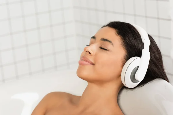 Mujer joven sonriente con los ojos cerrados descansando y escuchando música en los auriculares en el baño - foto de stock