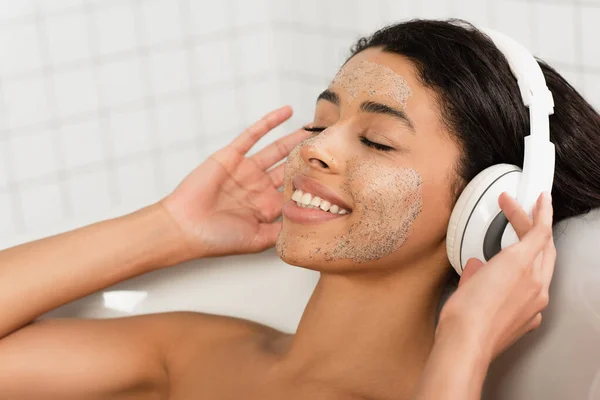Happy african american woman with scrub on face lying in bathtub with closed eyes and listening music in headphones — Stock Photo