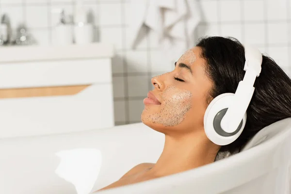 Lächelnde junge Frau mit geschlossenen Augen und Peeling auf dem Gesicht in Ruhe und Musik hören in Kopfhörern im Badezimmer — Stockfoto