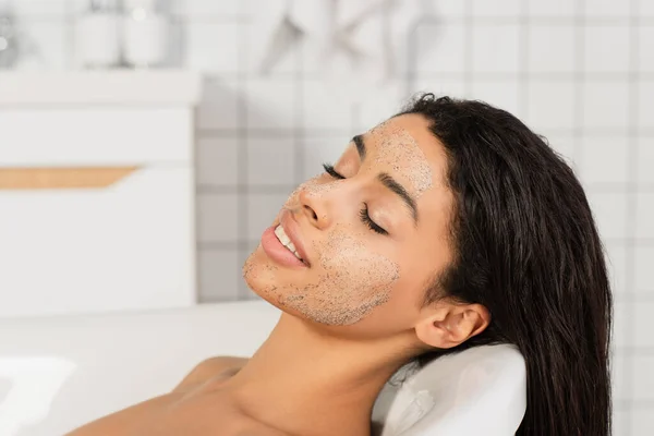 Mulher afro-americana com os olhos fechados e esfregar no rosto deitado na banheira — Fotografia de Stock