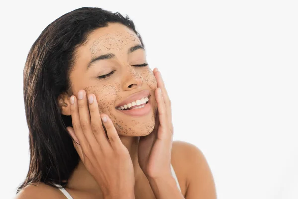 Cheerful young african american woman with closed eyes touching face with scrub isolated on white — Stock Photo