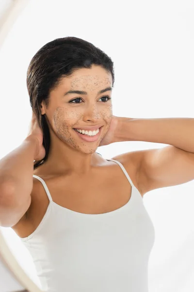 Afro-américaine avec gommage sur le visage regardant miroir et toucher les cheveux avec les mains sur fond blanc — Photo de stock