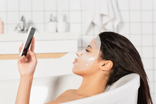 Mujer afroamericana joven con mascarilla en la cara tomando baño y sosteniendo el teléfono celular en el baño - foto de stock