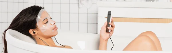 Femme afro-américaine avec masque en feuille sur le visage couché avec écouteurs et téléphone portable dans la baignoire, bannière — Photo de stock