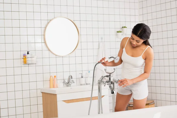 Young african american woman in white clothes preparing in bathtub and holding showerhead in hand in bathroom — Stock Photo