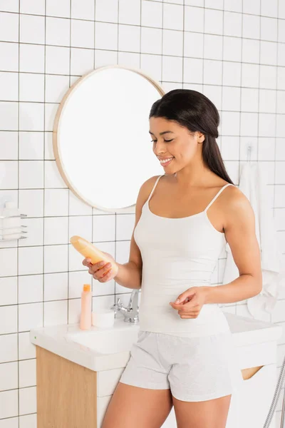 Sonriente joven afroamericana mujer en camiseta blanca y pantalones cortos sosteniendo gel de ducha en el baño - foto de stock