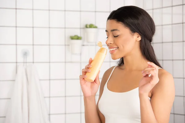 Smiling young african american woman in white tank top smelling aroma of shower gel in bathroom — Stock Photo