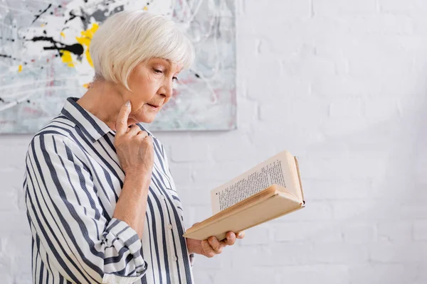 Старшая женщина в полосатой рубашке читает книгу дома — стоковое фото