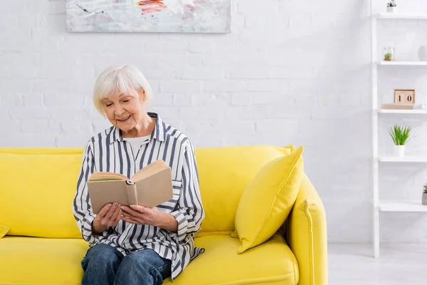 Lächelnde ältere Frau liest Buch, während sie auf der Couch sitzt — Stockfoto