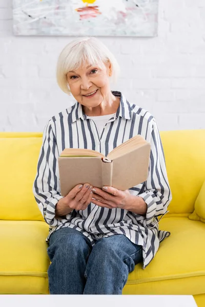 Seniorin mit Buch lächelt auf Couch in die Kamera — Stockfoto