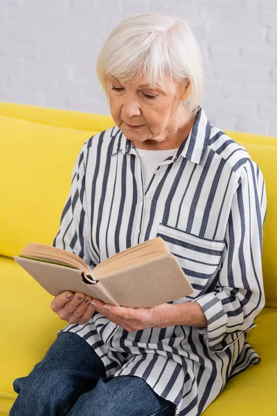 Ältere Frau liest Buch auf Couch im Wohnzimmer — Stockfoto