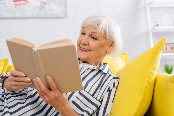 Happy elderly woman reading book on blurred foreground at home — Stock Photo