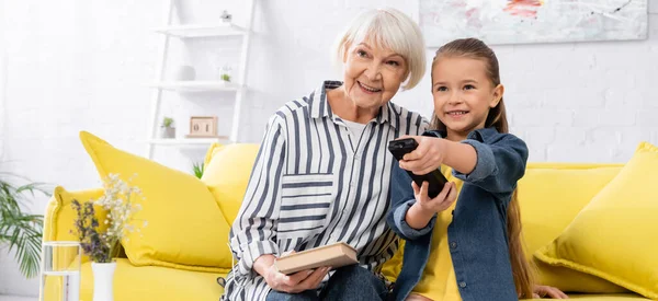 Smiling granny holding book near child clicking channels, banner — Stock Photo