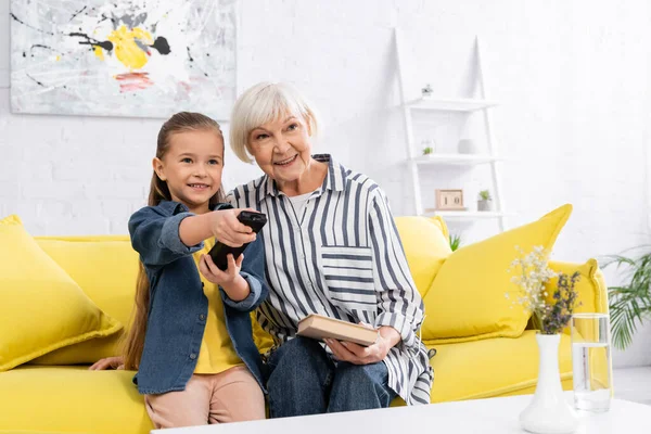 Bambino sorridente che tiene il telecomando vicino alla nonna con libro sul divano — Foto stock