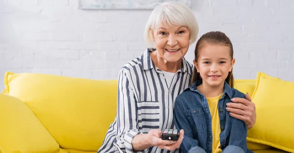 Souriant mamie étreignant enfant tout en regardant la télévision à la maison, bannière — Photo de stock