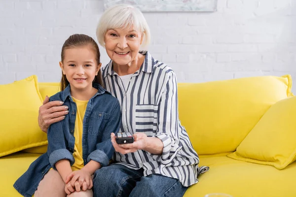 Grandmother with remote controller hugging child on couch — Stock Photo