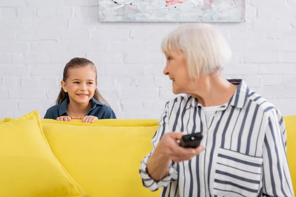 Child smiling at grandmother with remote controller on blurred foreground — Stock Photo