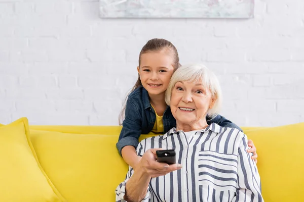 Enkelin umarmt Oma mit Fernbedienung zu Hause — Stockfoto