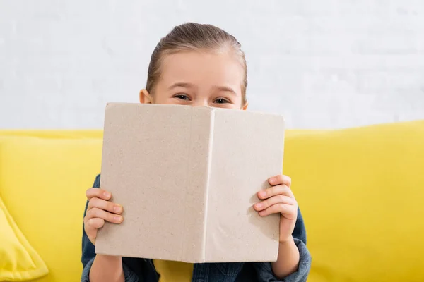 Child covering face with book on blurred couch — Stock Photo