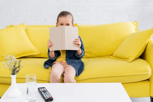 Menina segurando livro perto do rosto no sofá em casa — Fotografia de Stock