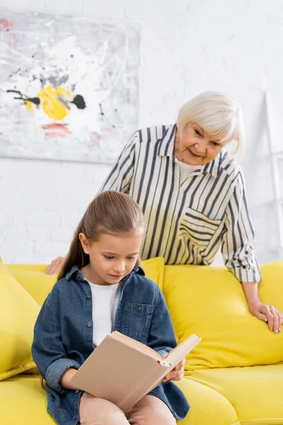 Дитина читає книгу біля розмитої бабусі вдома — стокове фото
